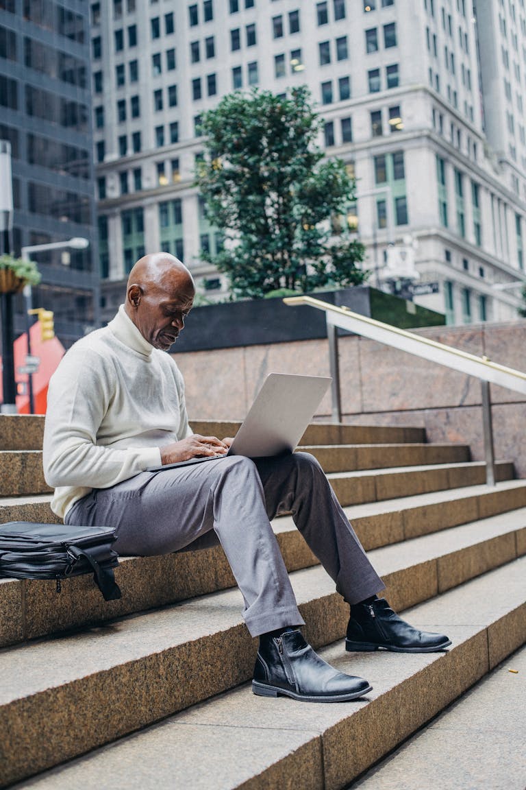 Full body of African American man in formal wear sitting on stairs in downtown and browsing netbook during work