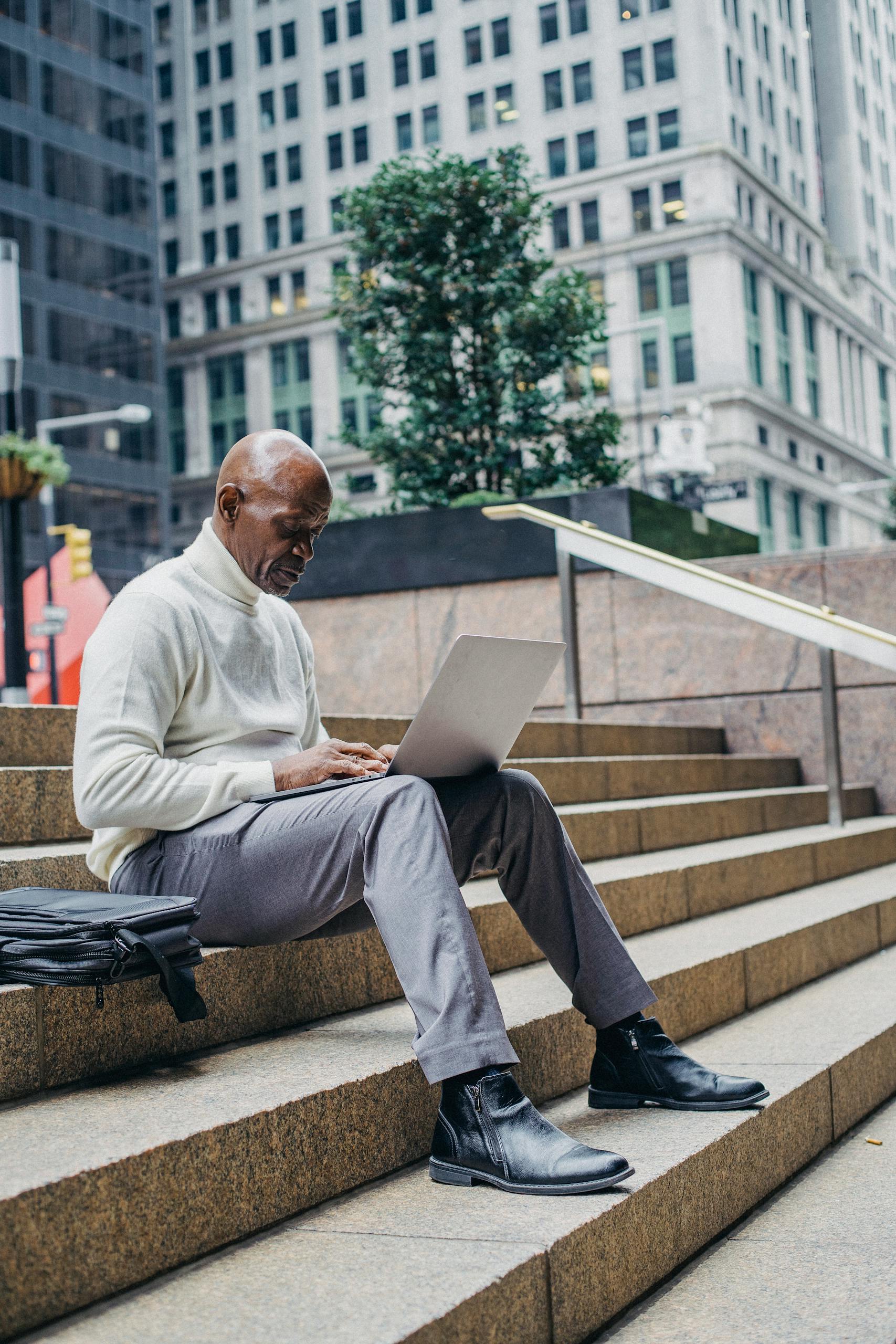 Full body of African American man in formal wear sitting on stairs in downtown and browsing netbook during work