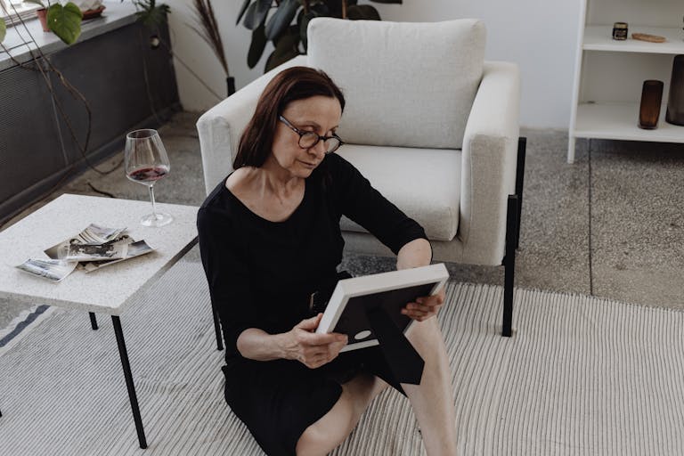 A mature woman sits on the floor in quiet reflection, holding a picture frame, a glass of wine nearby.