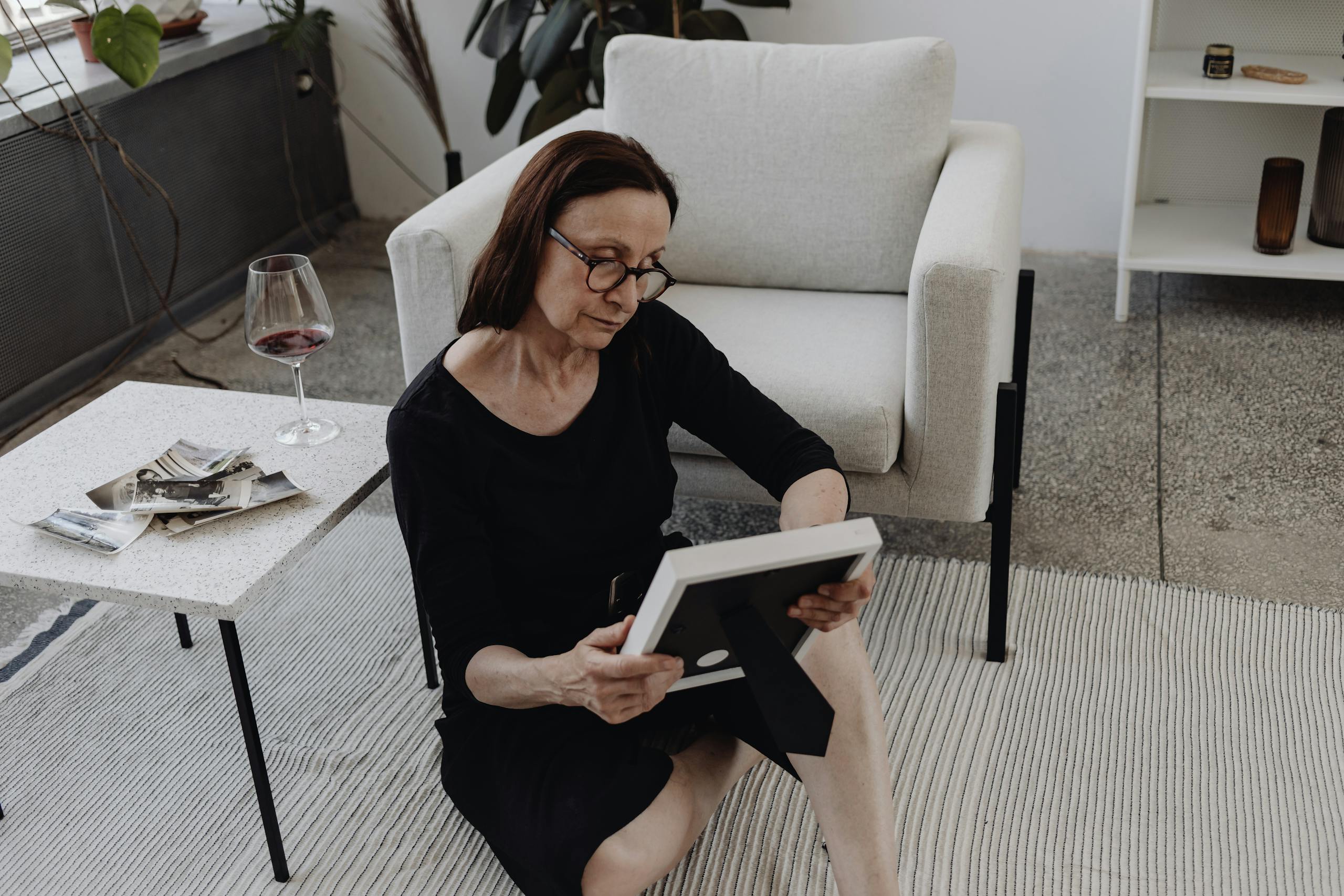 A mature woman sits on the floor in quiet reflection, holding a picture frame, a glass of wine nearby.