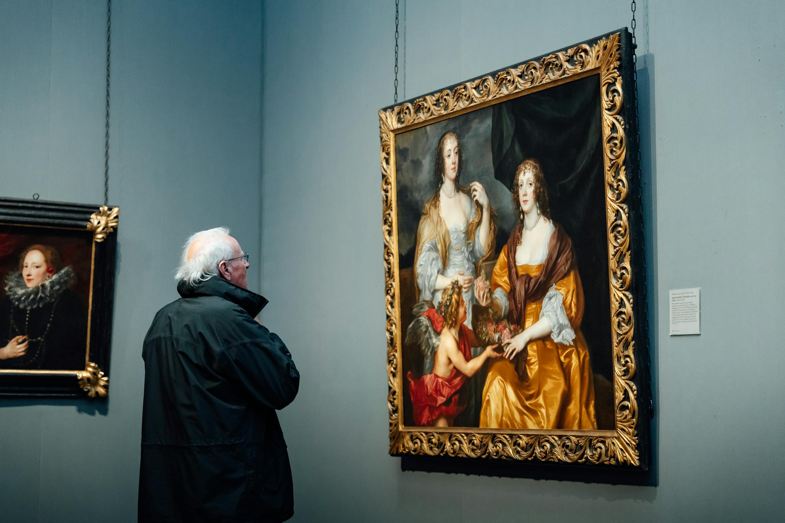 A senior man observes classical paintings in an art museum gallery.