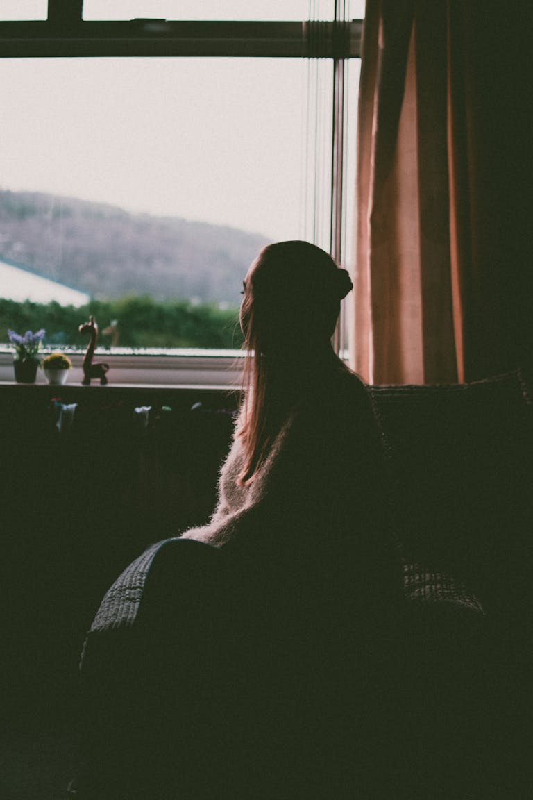 A silhouette of a woman sitting by a window, gazing outside during daytime.