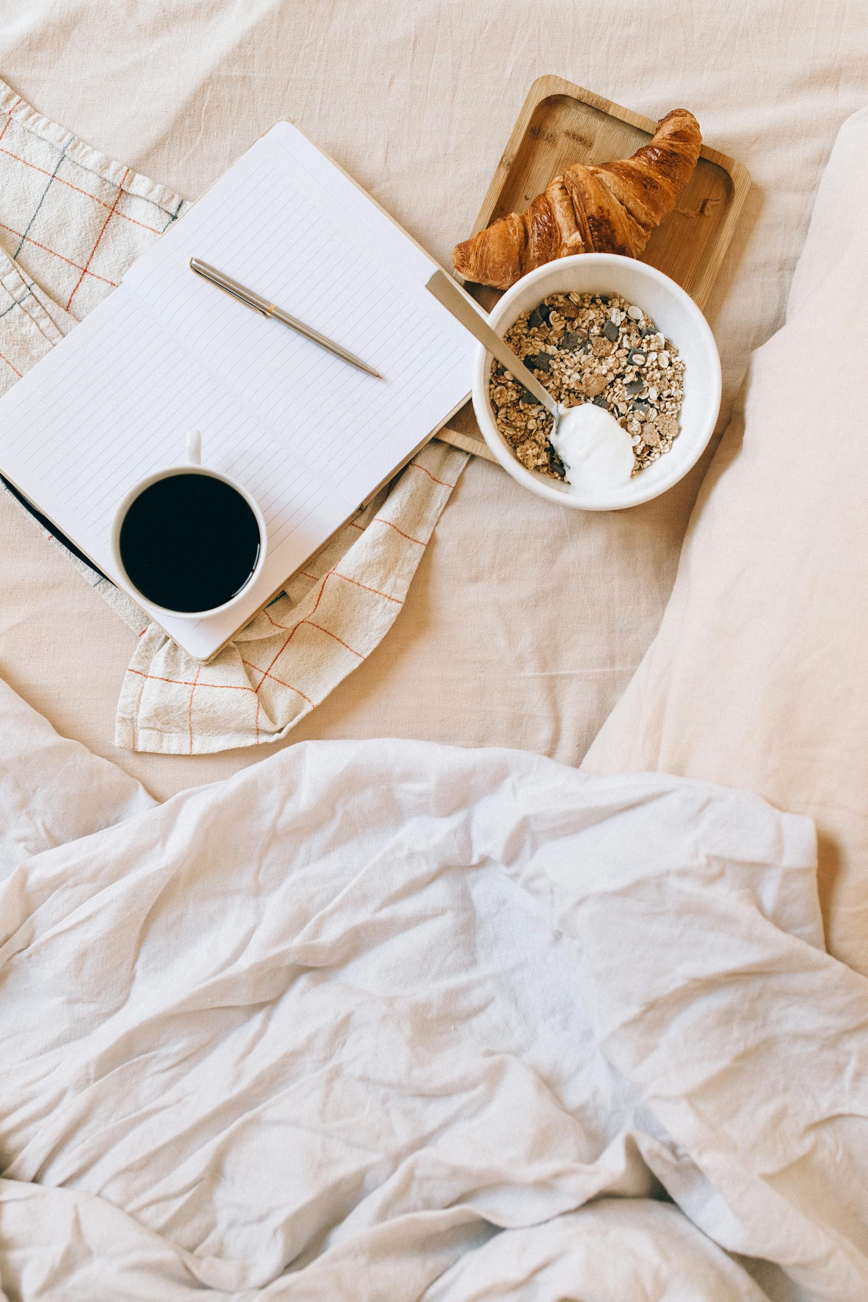 A warm and inviting breakfast setup with coffee, croissant, and yogurt granola bowl on a comfy bed.