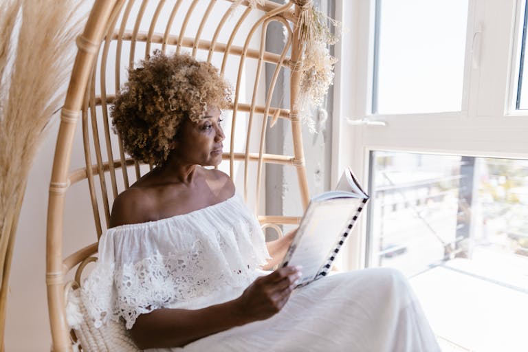 A woman with curly hair reads in a wicker chair beside a sunlit window, embracing tranquility.