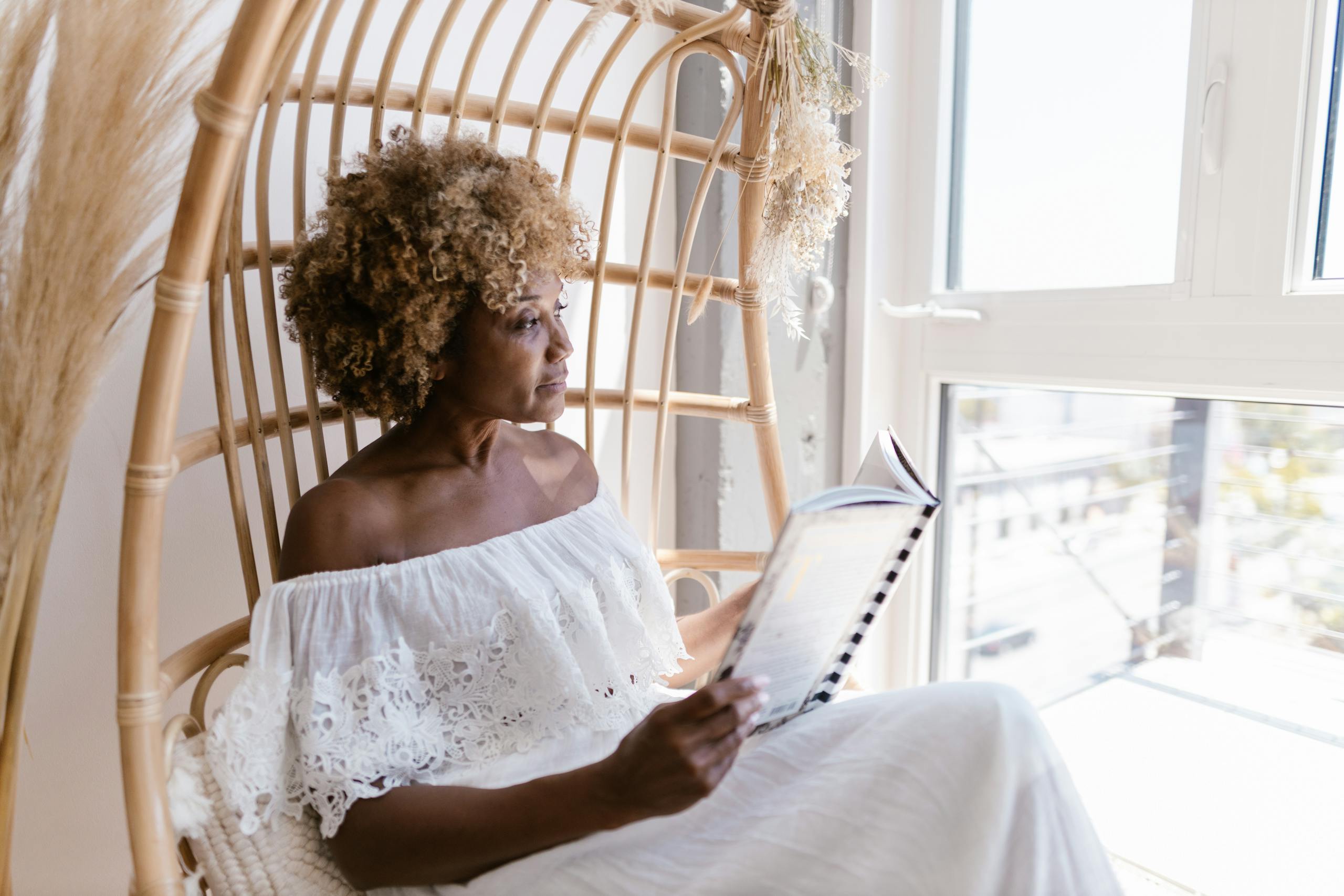 A woman with curly hair reads in a wicker chair beside a sunlit window, embracing tranquility.