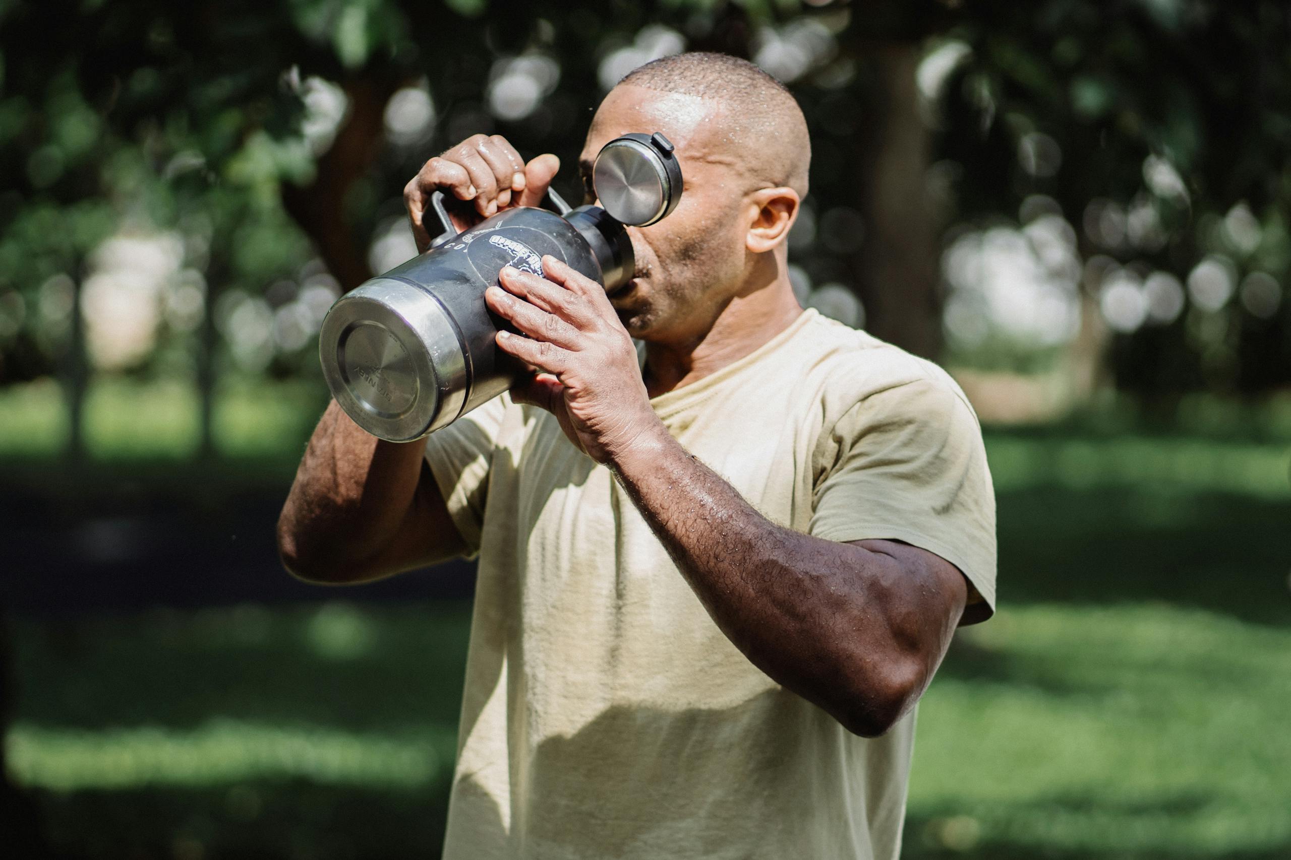 An adult man drinking from a large tumbler outdoors in a green park setting.