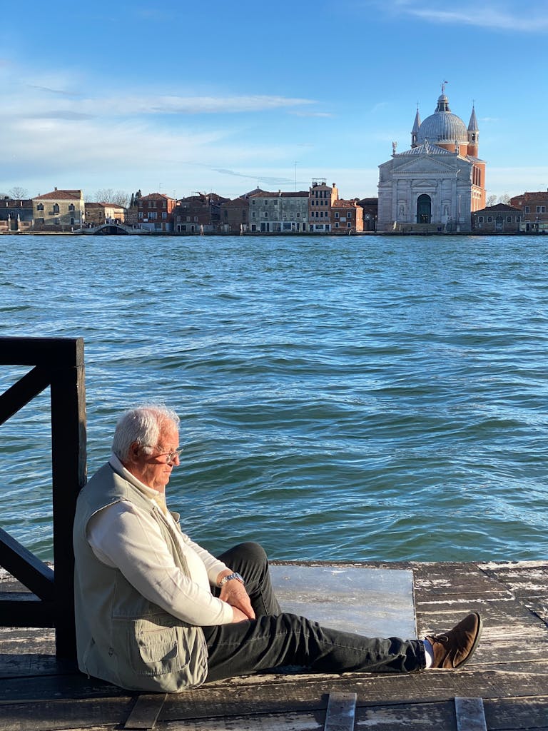An elderly man sits by the canal in Venice, enjoying a tranquil moment against a scenic backdrop.