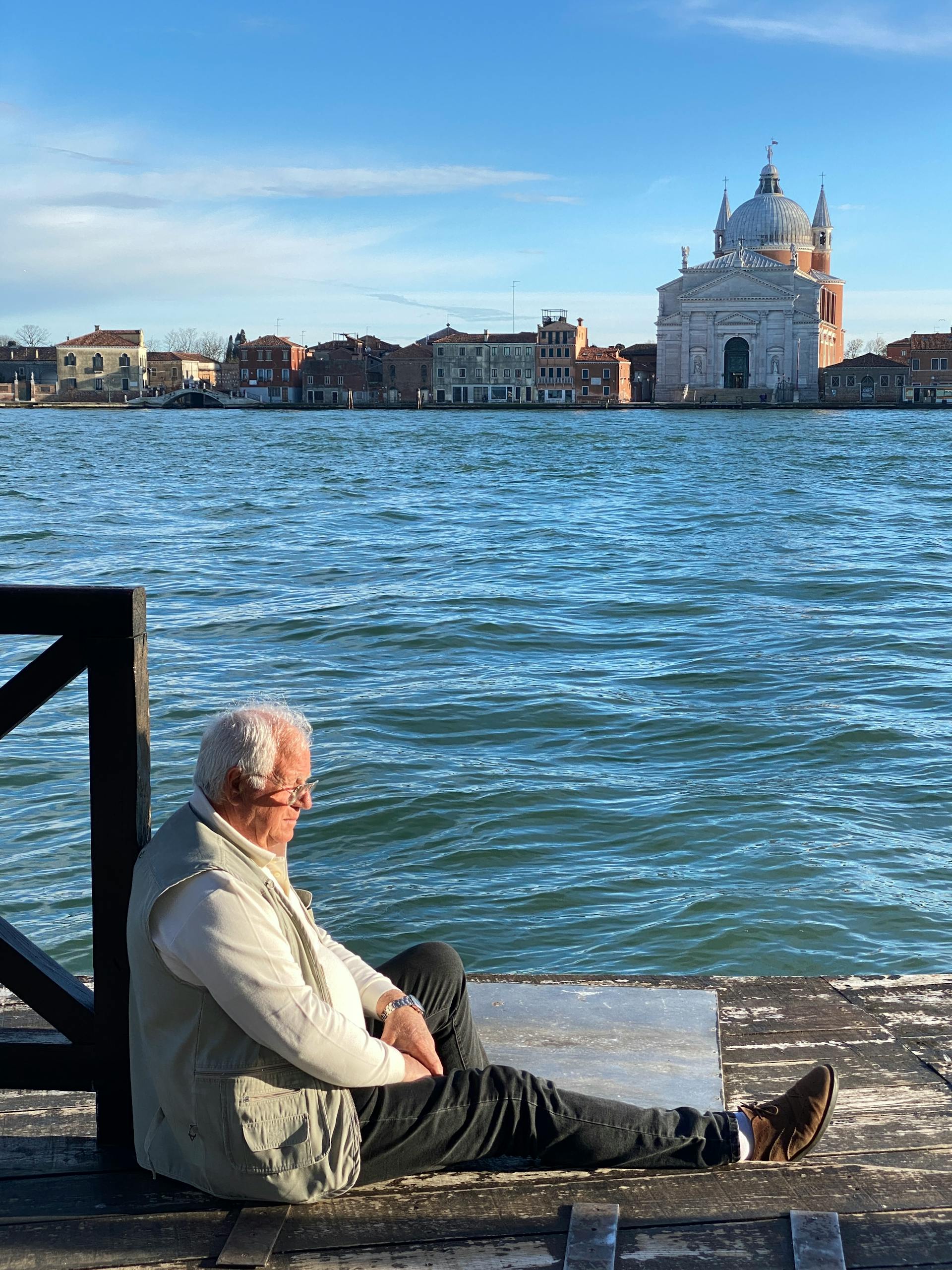 An elderly man sits by the canal in Venice, enjoying a tranquil moment against a scenic backdrop.