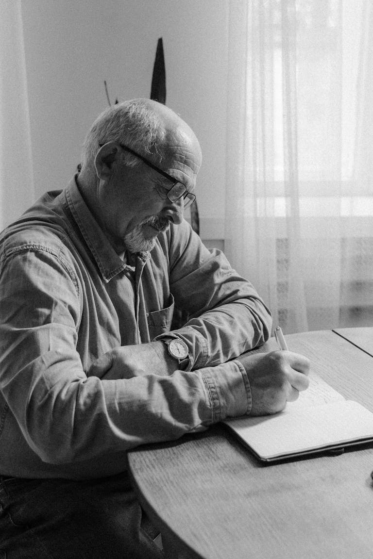Black and white photo of a senior man writing in a notebook at a table indoors.