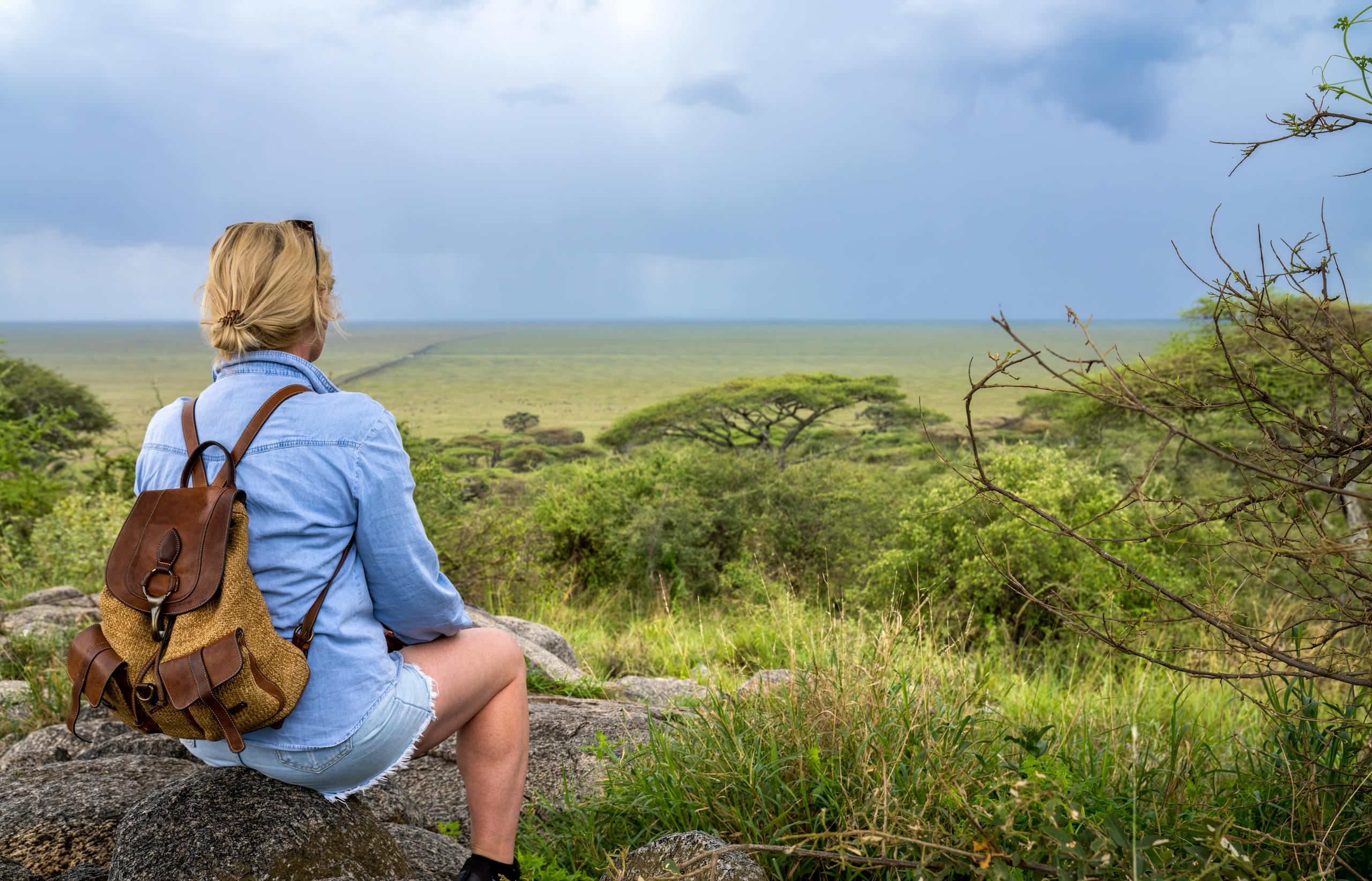 Blonde woman with backpack enjoying view of expansive African landscape under overcast skies.