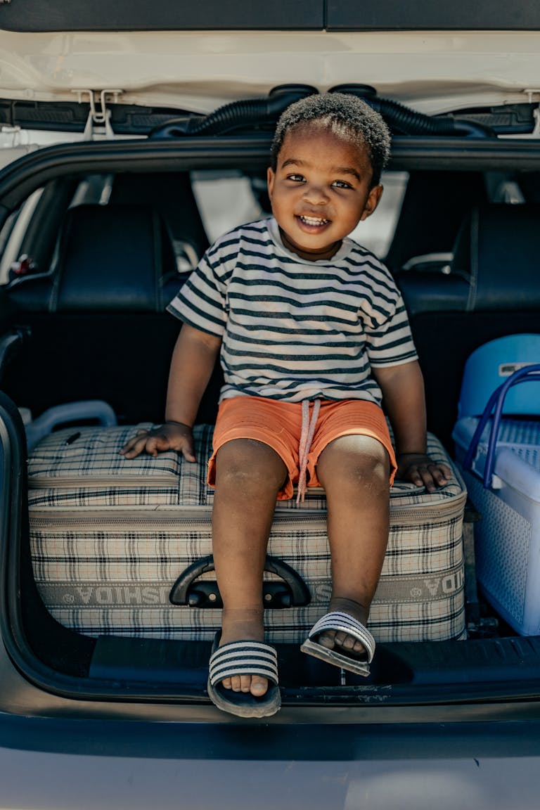 Cheerful child in a car trunk sitting on a suitcase, ready for a travel adventure.