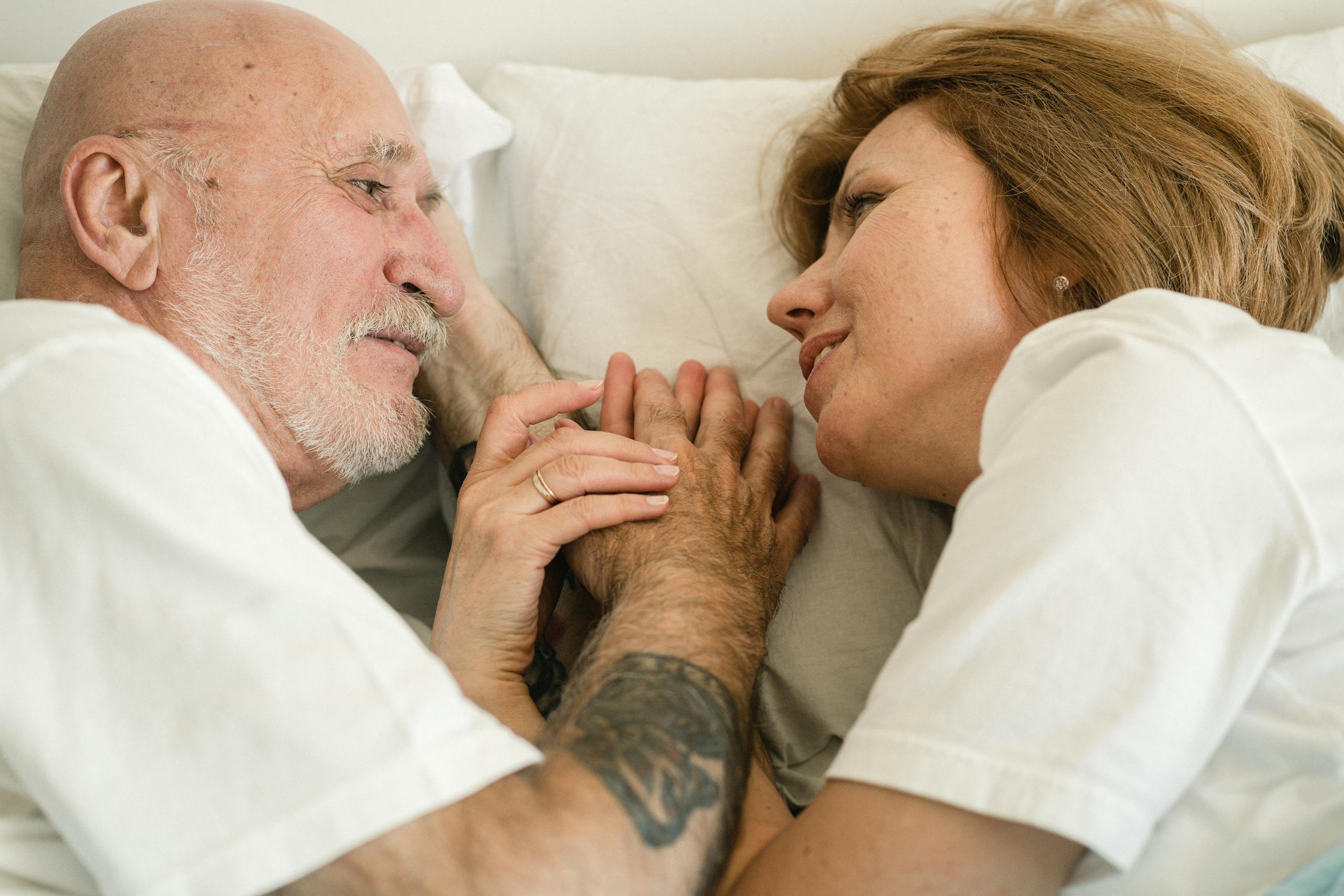 Close-up of a senior couple holding hands and smiling at each other while lying in bed, showcasing love and connection.