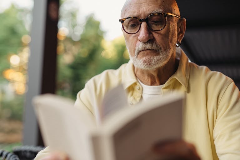Elderly man with eyeglasses focused on reading a book outside.