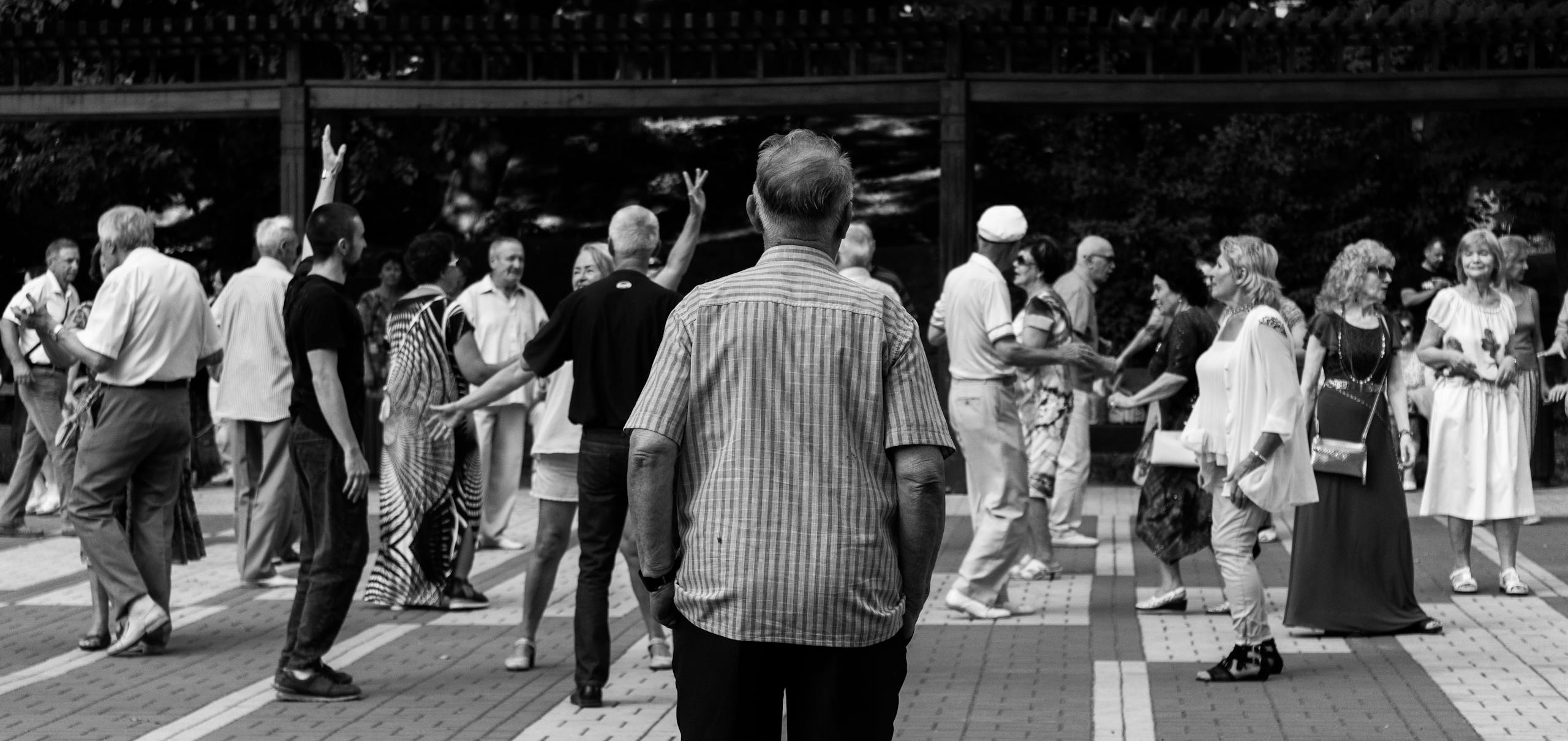 Elderly people socializing and dancing outdoors in a lively urban gathering captured in black and white.