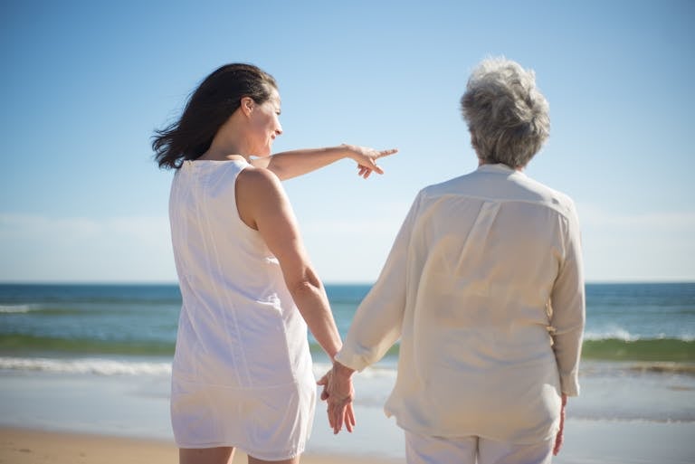 Elderly woman and adult daughter holding hands on a sunny beach in Portugal.
