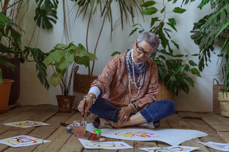 Elderly woman painting watercolors surrounded by plants indoors, artistically focused.