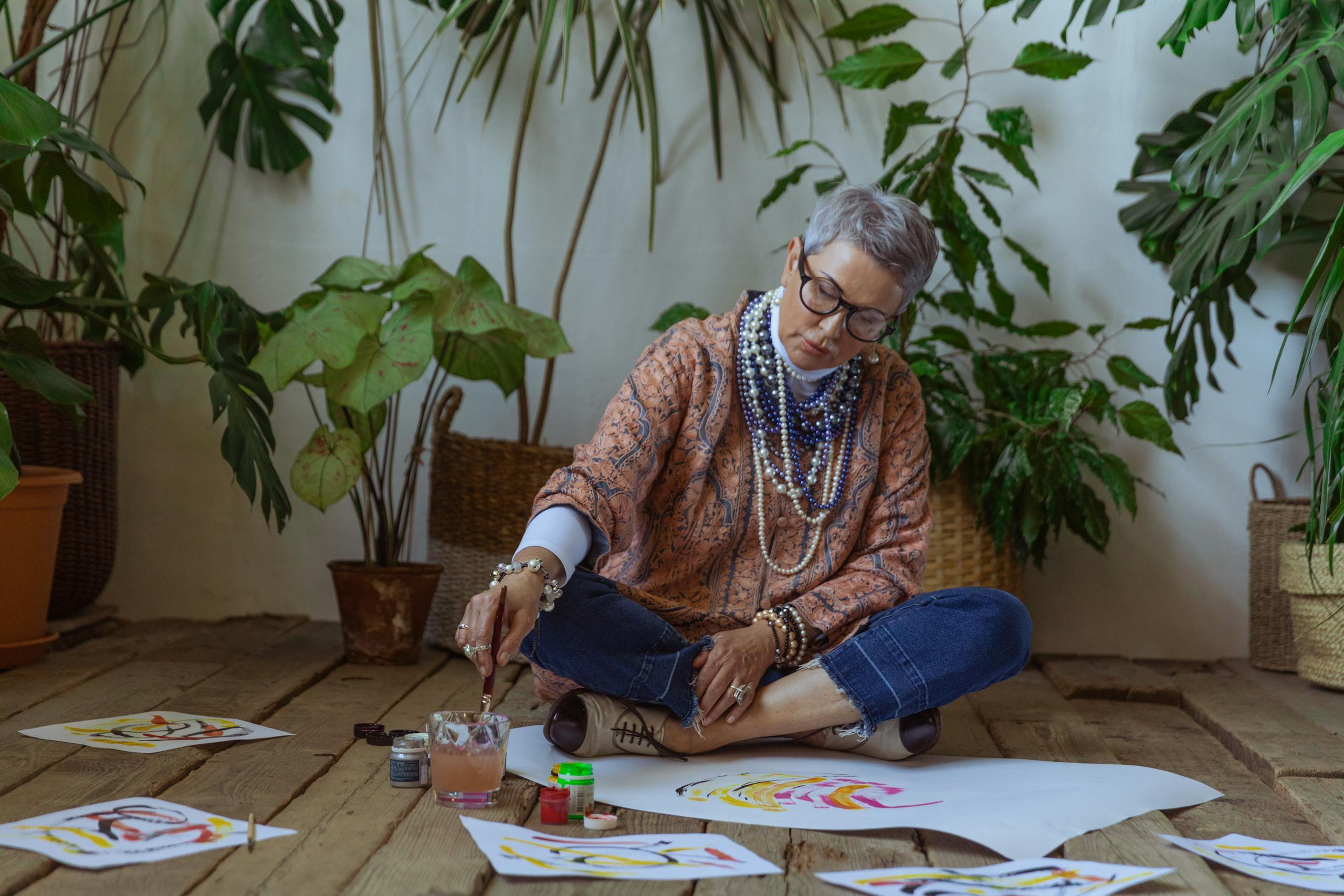 Elderly woman painting watercolors surrounded by plants indoors, artistically focused.