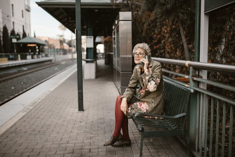 Elderly woman sitting on bench at outdoor train station using smartphone.