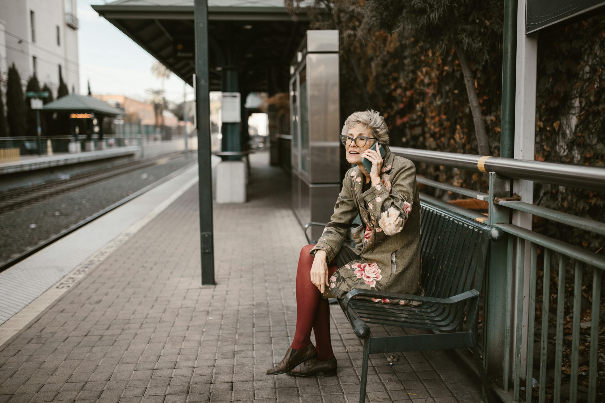 Elderly woman sitting on bench at outdoor train station using smartphone.