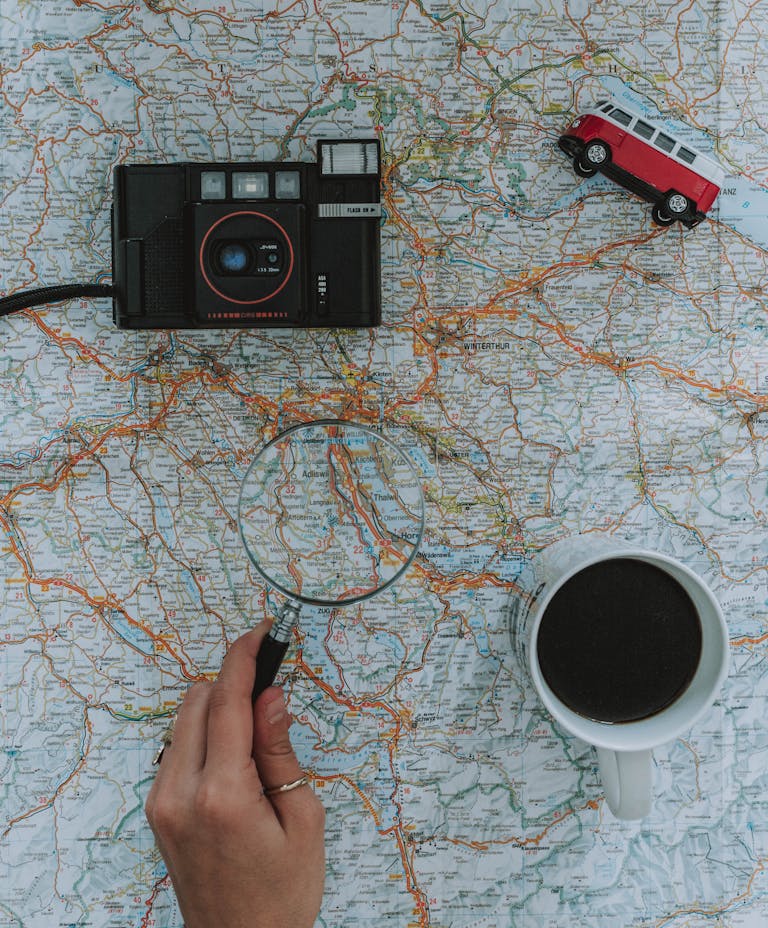 Flat lay of travel essentials: map, camera, toy van, magnifying glass, and coffee.