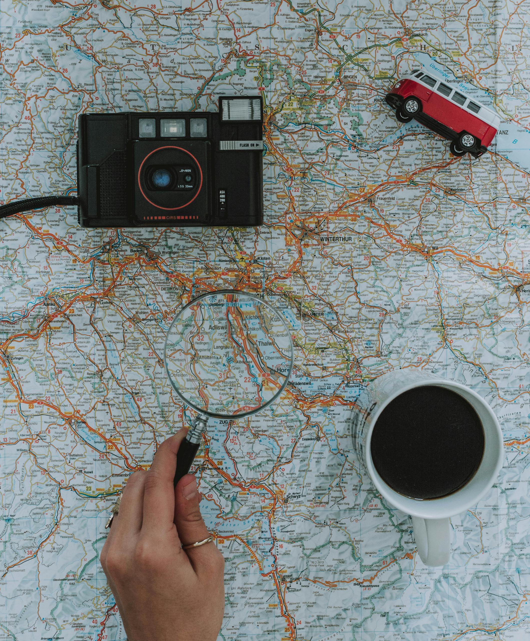 Flat lay of travel essentials: map, camera, toy van, magnifying glass, and coffee.