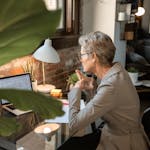 Senior woman in a modern home office focusing on work at a laptop next to a brick wall.
