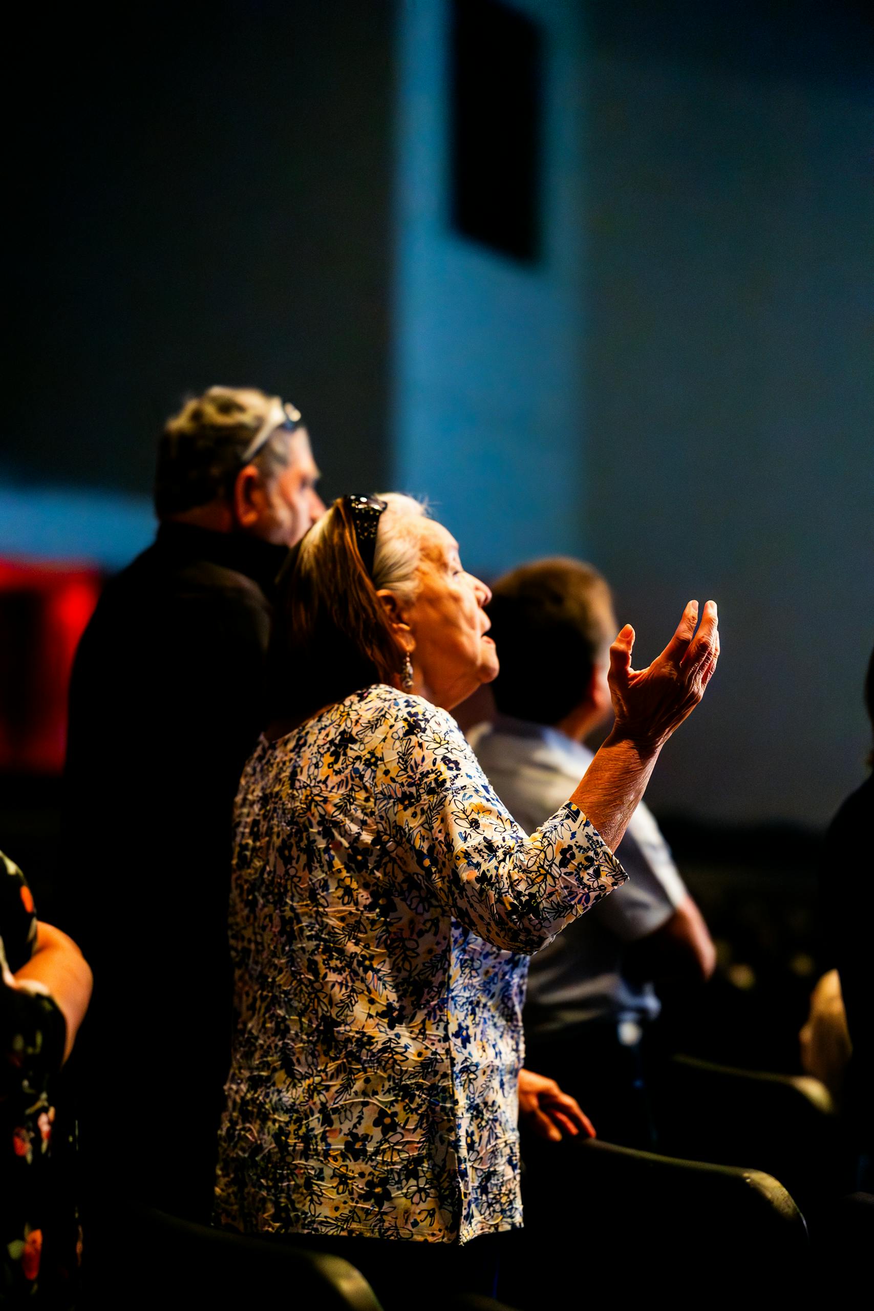 Senior woman with arm raised in prayer during a group meeting inside a dimly-lit hall.