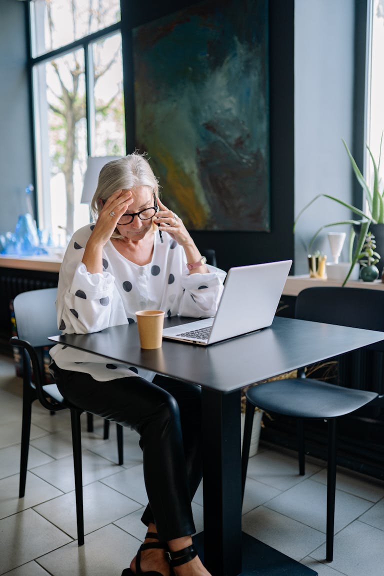 Senior woman with white hair looks stressed while working on a laptop in a modern office.