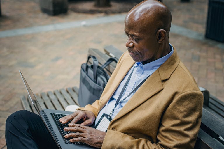 Side view crop serious African American male in formal wear sitting on bench and typing on netbook