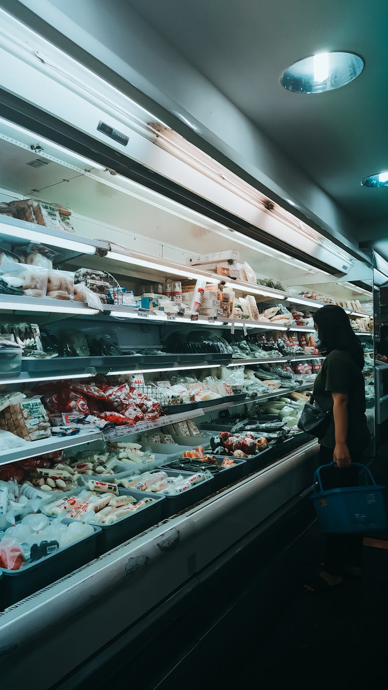 Side view of unrecognizable young female in casual clothes and mask with basket in hand standing near fridge showcase and choosing various products in supermarket