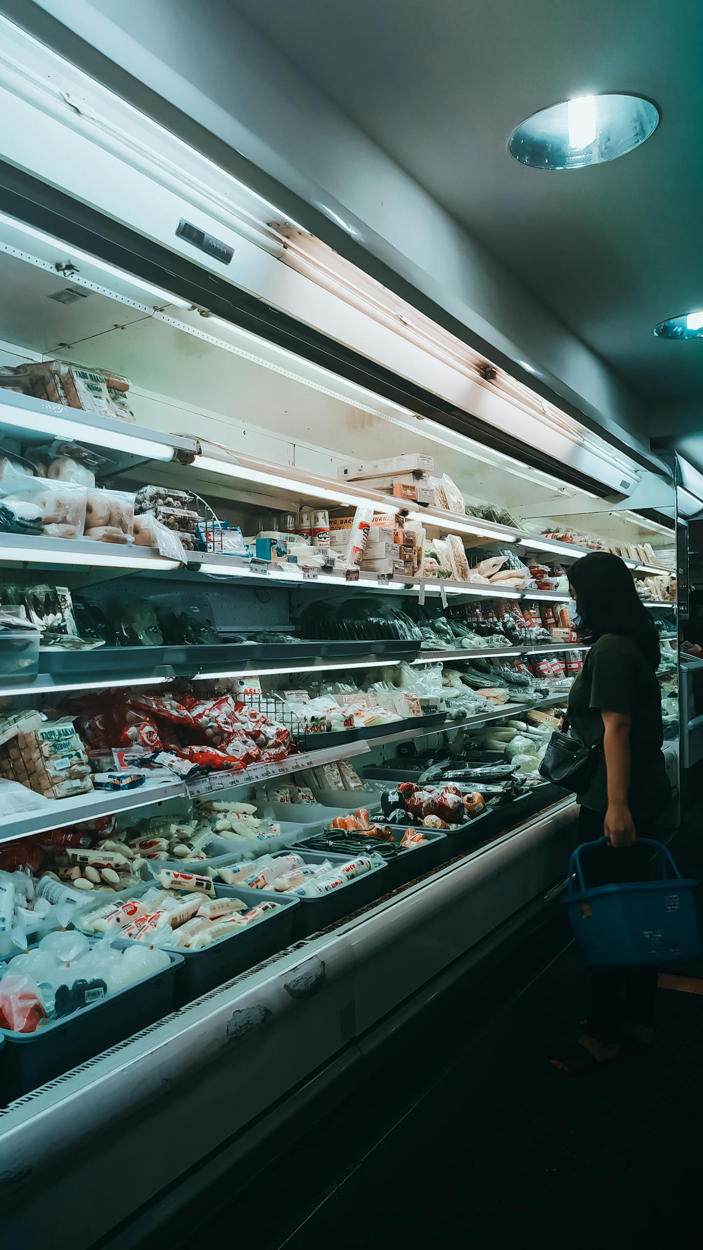 Side view of unrecognizable young female in casual clothes and mask with basket in hand standing near fridge showcase and choosing various products in supermarket