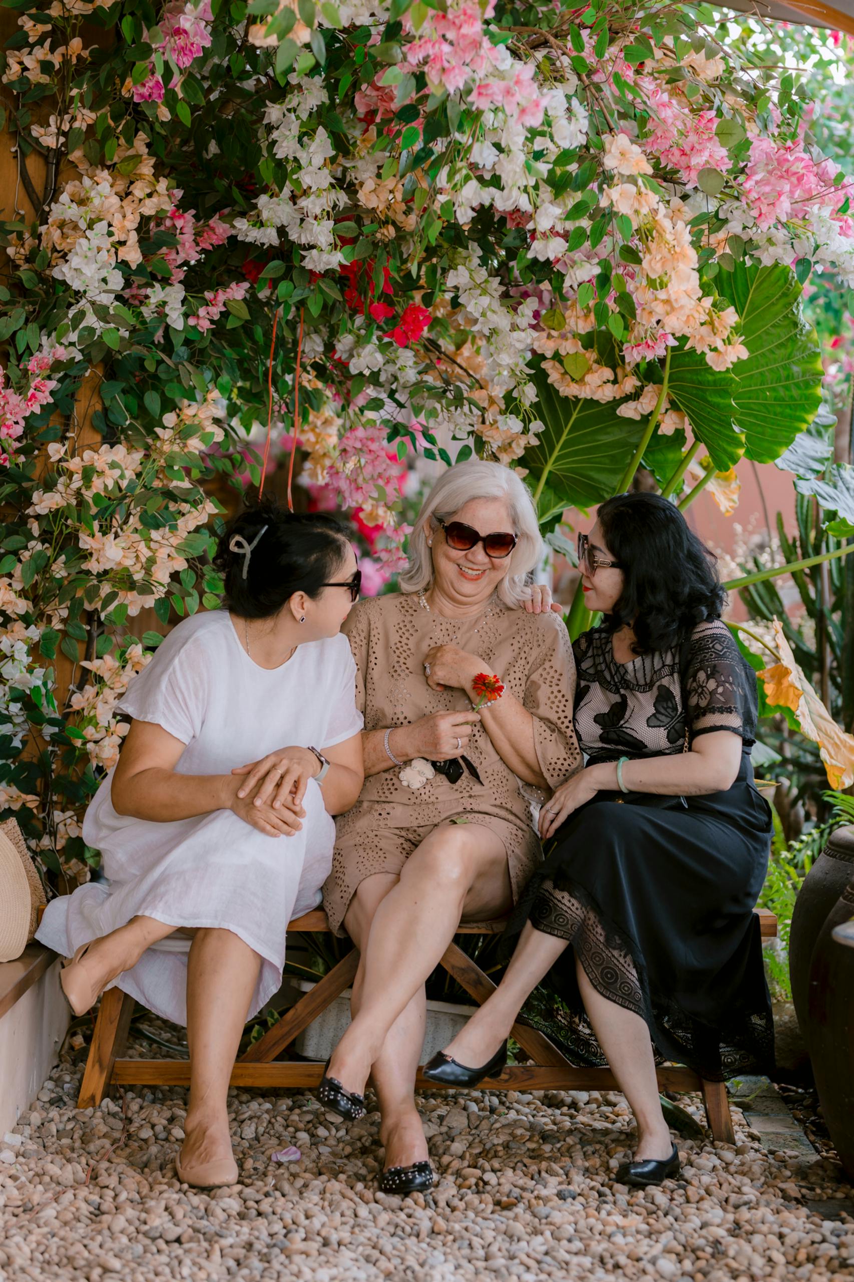 Three women sitting outdoors surrounded by vibrant flowers, enjoying a joyful conversation.