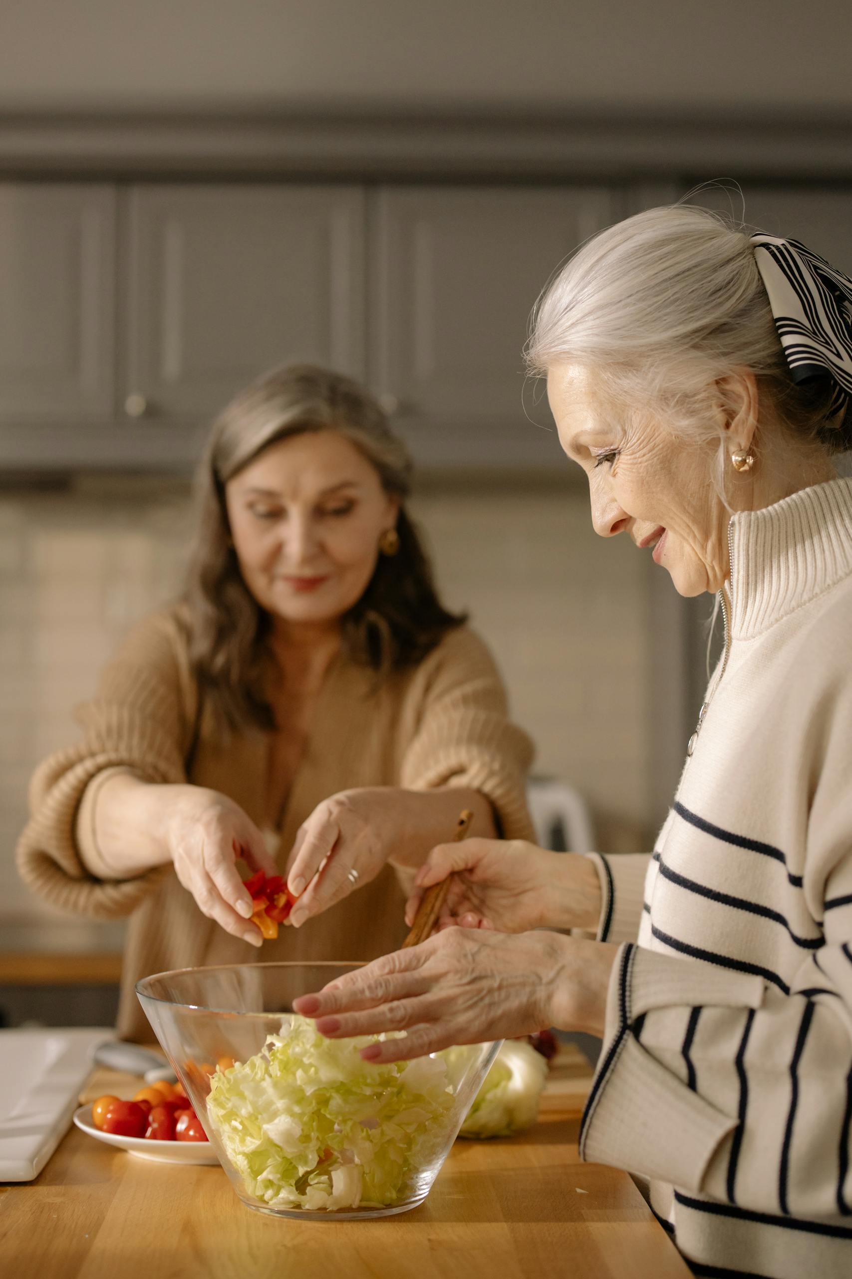 Two elderly women preparing a fresh salad with cherry tomatoes in a cozy kitchen setting.