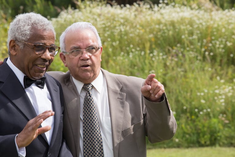 Two senior men sharing a joyful moment in a sunny outdoor setting, dressed formally in suits.