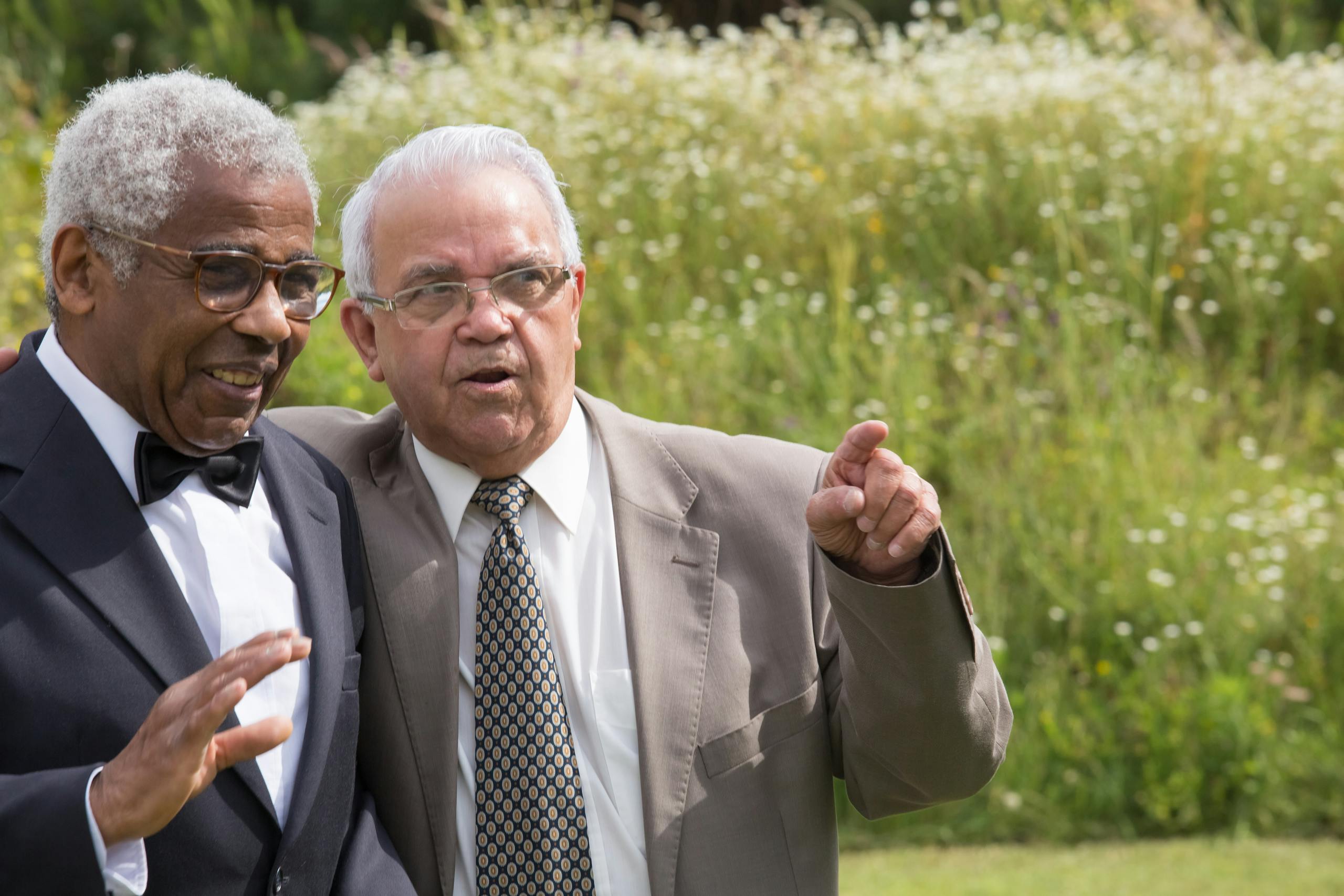 Two senior men sharing a joyful moment in a sunny outdoor setting, dressed formally in suits.