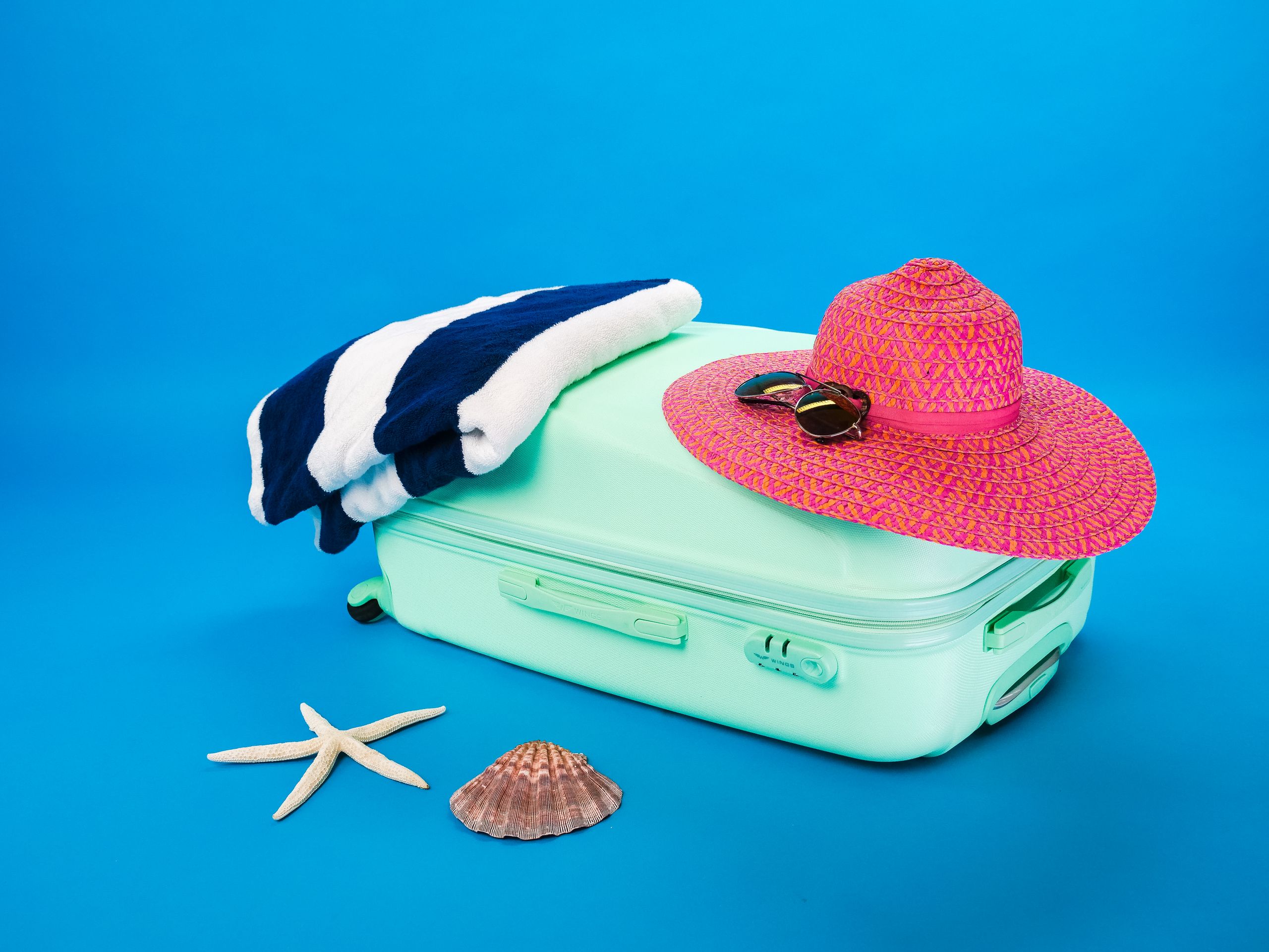 Beach essentials including a suitcase, hat, towel, and seashells on a bright background.