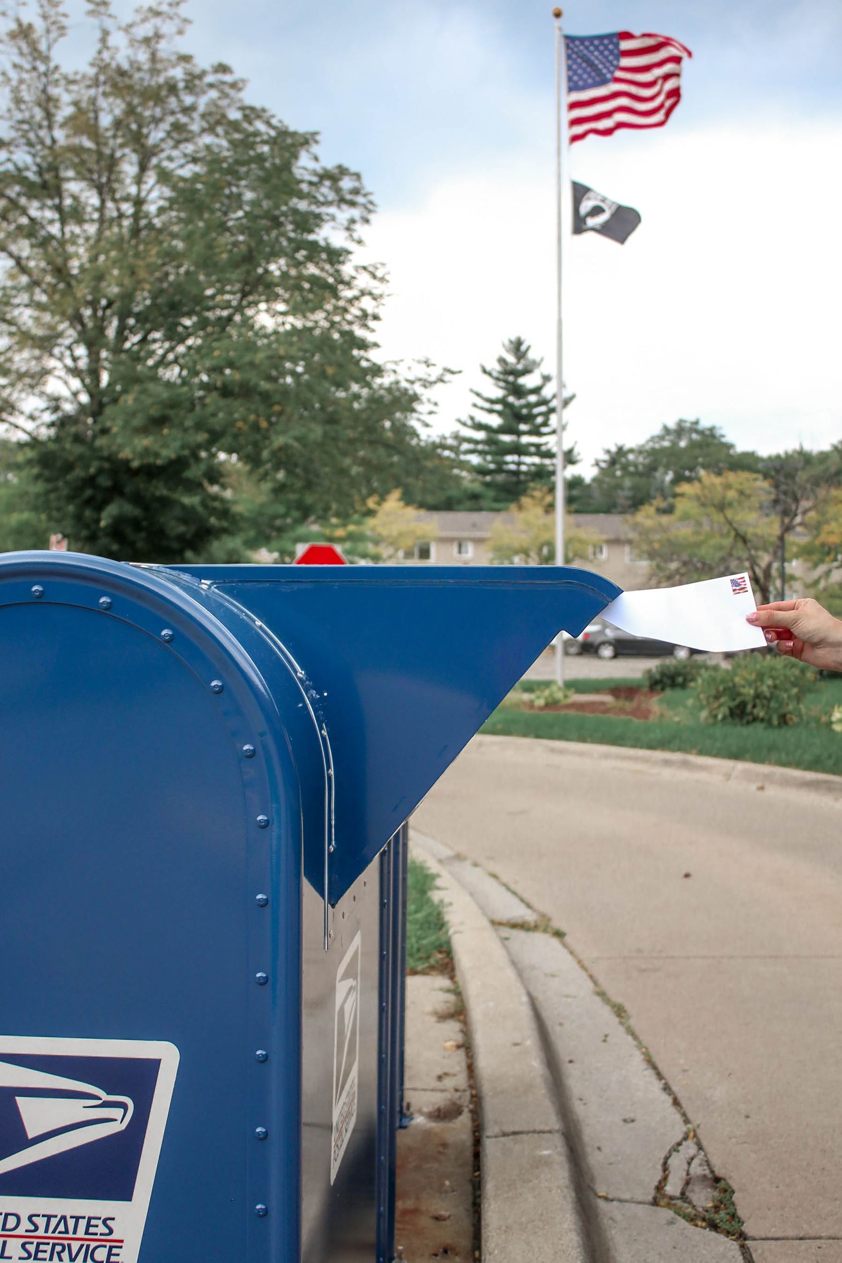 Crop unrecognizable person putting letter in outdoor blue metal mailbox against waving flag of United States