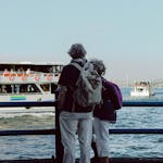 Elderly couple stands by the waterfront watching boats on a sunny day.