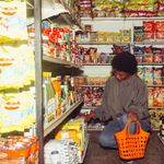 Full length of adult ethnic female buyer selecting goods while squatting with shopping bag among grocery shelves