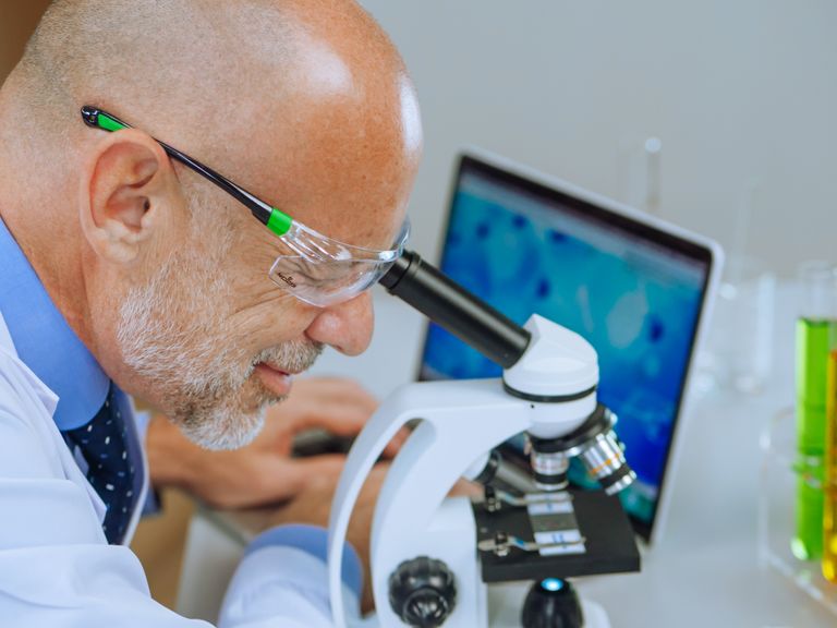 Senior scientist in lab coat using microscope, focused on research with laptop in background.
