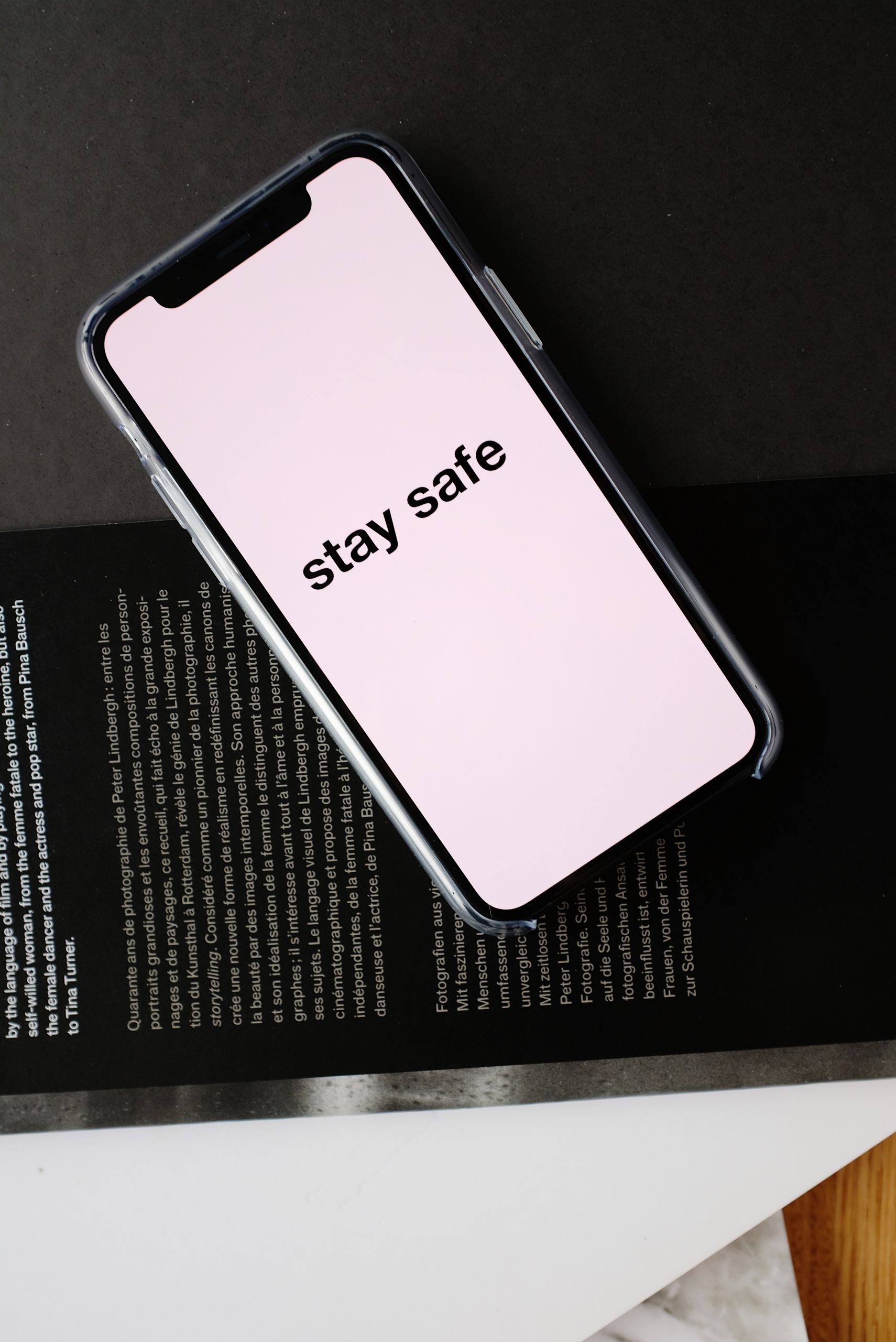 Top view of modern mobile phone with pale pink screen and STAY SAFE inscription placed on open magazine with black page and white letters during COVID 19 pandemic