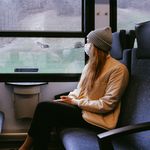 Woman wearing mask on train during pandemic, observing outdoor scenery.