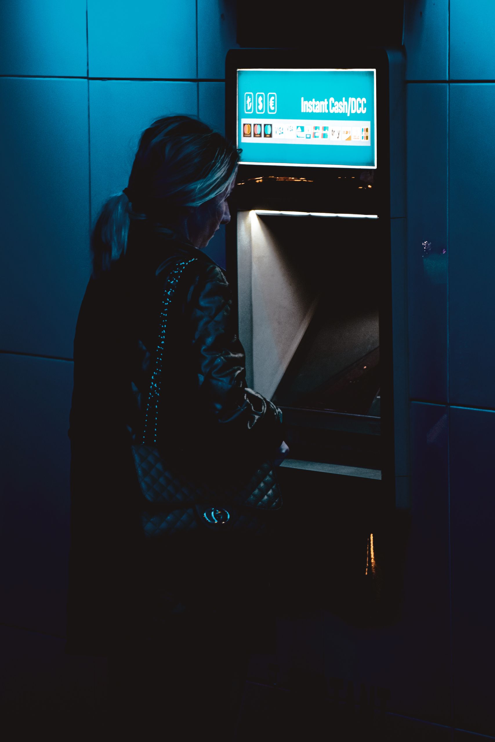 A woman uses an ATM at night in Istanbul under moody blue lighting.