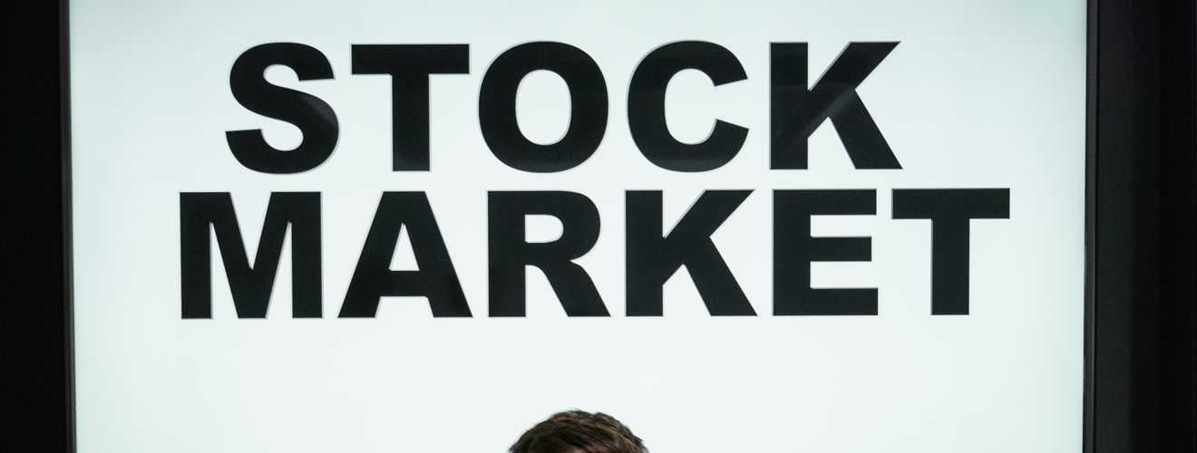 Professional businessman with arms crossed in front of a stock market display in a modern office setting.