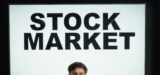 Professional businessman with arms crossed in front of a stock market display in a modern office setting.