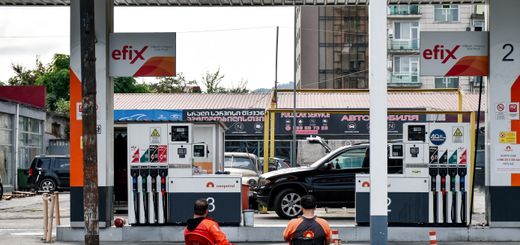 Two men sitting at a gas station with fuel pumps and cityscape background.