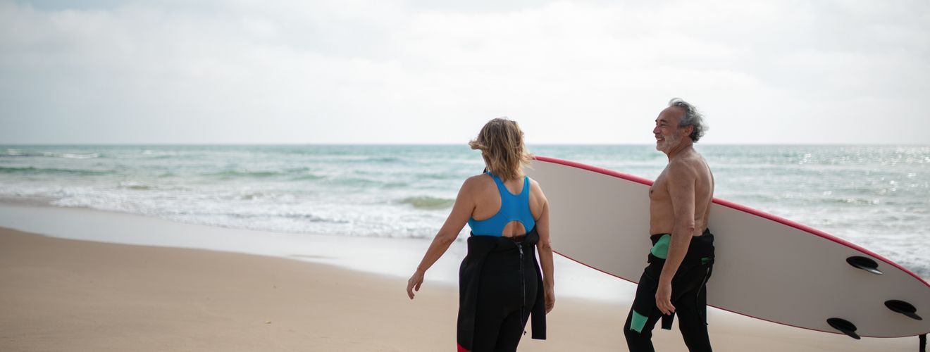 Two senior surfers on a beach in Portugal, with a surfboard and the ocean waves in the background.
