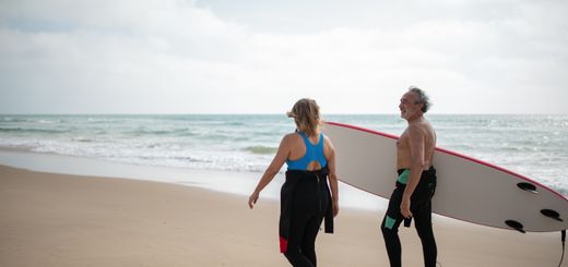 Two senior surfers on a beach in Portugal, with a surfboard and the ocean waves in the background.