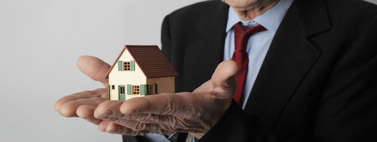 Elderly man in business attire holding a small house model, symbolizing real estate investment.