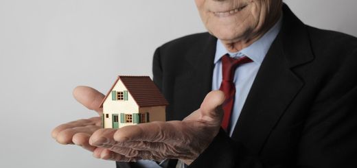 Elderly man in business attire holding a small house model, symbolizing real estate investment.